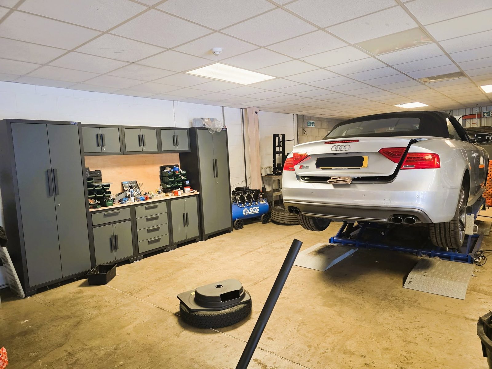 Silver Audi S5 convertible raised on a lift beside grey workshop cabinets